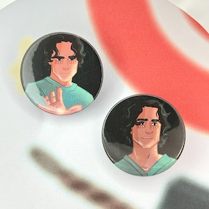 May include: Two round buttons featuring a stylized illustration of a person with dark curly hair and a teal shirt. The person is depicted with a serious expression. The background of the buttons is black.