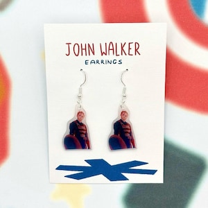 May include: A pair of dangle earrings featuring a character in a blue and red uniform with a shield. The earrings are on a white card with the text "JOHN WALKER EARRINGS" in red. The background has a colorful pattern.