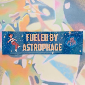 May include: A blue sticker with gold text reading "FUELED BY ASTROPHAGE". The sticker features an illustration of a person in a white shirt and red trousers, and a geometric shape. The background is a starry sky.