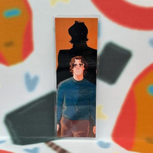 May include: A bookmark featuring an illustration of a person in a blue sweater and brown pants, with a black silhouette behind them. The background is orange, and the bookmark is in a clear plastic sleeve.