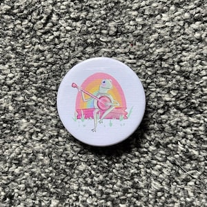 May include: A white button featuring a cartoon frog playing a banjo in front of a rainbow. The frog is green with a light blue eye and is sitting on a brown log.