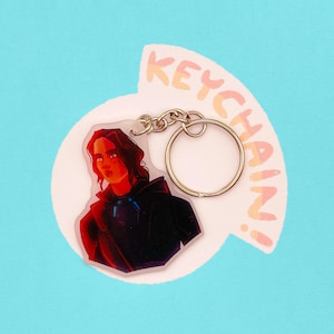 Può includere: Un portachiavi in acrilico trasparente con un anello e una catena in metallo argentato. Presenta un'illustrazione di una persona con una giacca con cappuccio nera. La parola "KEYCHAIN!" è scritta a semicerchio sopra.