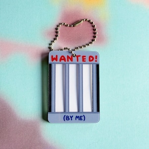 May include: A rectangular keyring featuring a jail cell design with vertical bars. The top text reads WANTED in red, and the bottom text reads (BY ME) in blue. The keyring is attached to a silver-coloured metal ball chain for hanging.