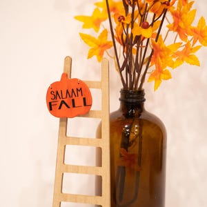 May include: A small wooden ladder with a pumpkin-shaped sign that says "Salaam Fall" hanging from the top rung. A brown glass vase with a bouquet of orange and yellow fall leaves sits next to the ladder.