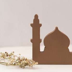 May include: A brown wooden cutout of a mosque with a single minaret. The mosque is on a white surface with a sprig of dried white flowers in the foreground.