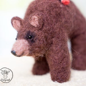 May include: A brown needle felted bear with black eyes and a pink nose. The bear is standing on a white surface.