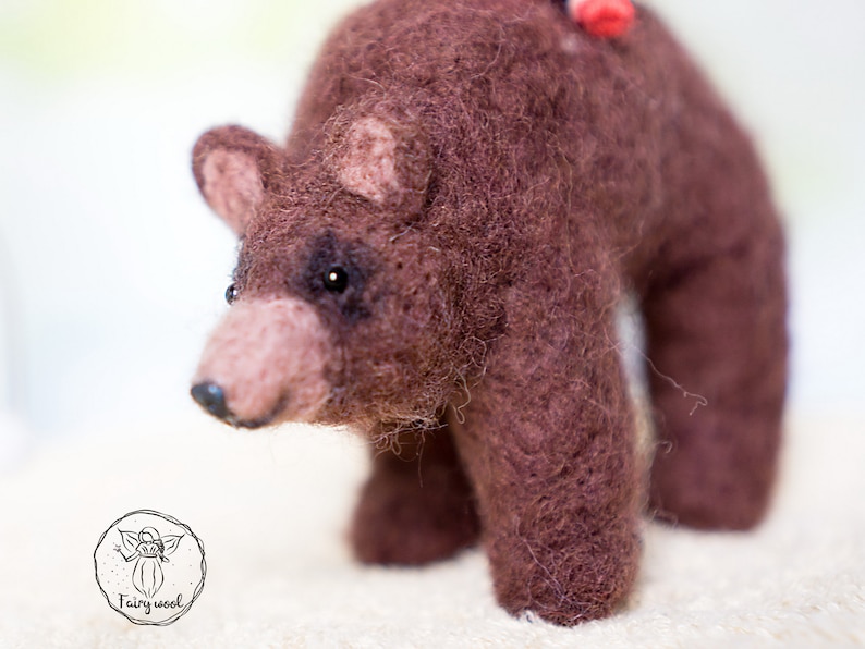 May include: A brown needle felted bear with black eyes and a pink nose. The bear is standing on a white surface.