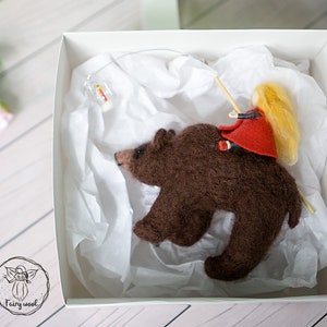 May include: A brown felt bear with a girl in a red dress and blonde hair riding on its back. The bear and girl are in a white box with white tissue paper.