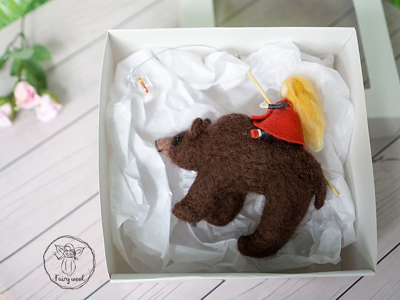 May include: A brown felt bear with a girl in a red dress and blonde hair riding on its back. The bear and girl are in a white box with white tissue paper.
