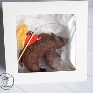 May include: A small, brown felt bear with a girl riding on its back. The girl is wearing a red dress and has long blonde hair. The bear and girl are in a white box with a clear plastic window.