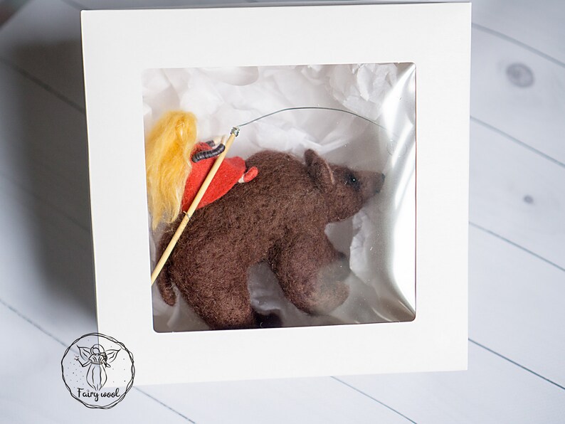 May include: A small, brown felt bear with a girl riding on its back. The girl is wearing a red dress and has long blonde hair. The bear and girl are in a white box with a clear plastic window.