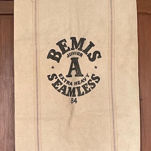 Puede incluir: Un saco de arpillera blanco vintage con texto impreso en negro que dice "Bemis Junior A Extra Heavy Seamless 84".