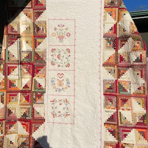 Op de afbeelding: Een wit quilt met een blokhutpatroon aan de ene kant en geborduurde bloemenontwerpen aan de andere kant. De bloemenontwerpen omvatten een vaas met bloemen, een krans, een hart en een vogel.