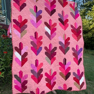 Op de afbeelding: Een roze quilt met een geometrisch patroon van overlappende bladeren in tinten roze, rood, paars en bruin. De quilt is gemaakt van verschillende stoffen en texturen.