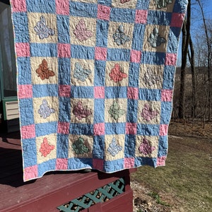 May include: A vintage quilt with a patchwork design featuring a grid of blue, pink, and cream squares. Each square has a butterfly applique in a variety of colors, including brown, pink, green, and blue.