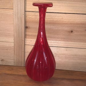 Può includere: Un vaso alto di vetro rosso a forma di goccia e con collo a spirale. Il vaso ha una parte superiore piatta e rettangolare e scanalature verticali lungo il corpo. Il vaso è alto circa 30 cm.