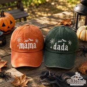 May include: Two baseball caps, one orange and one dark green, each featuring white embroidered text that reads 'mama' or 'dada' in a gothic font, accented with white spiderweb, spider, and bat motifs on the front panels of the hats.