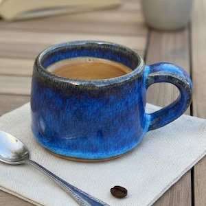 May include: A blue ceramic coffee mug filled with coffee sits on a beige linen napkin, next to a silver spoon and a coffee bean. An open book and a small potted succulent are in the background. The mug has a dark blue rim and handle.
