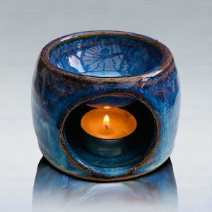 May include: A blue ceramic oil burner with a teal glaze and a brown rim. A small tea light candle is burning inside the burner.