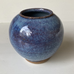 May include: A small, round, blue ceramic vase with a speckled glaze. The vase has a slightly wider opening at the top and a small, rounded base.