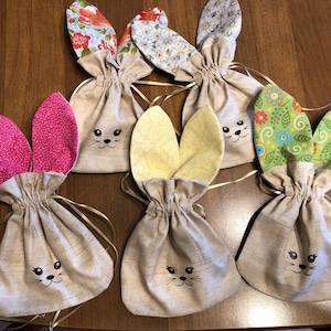 May include: Six handmade bunny-shaped fabric gift bags with different patterned ears. The bags are made of natural linen fabric and have a drawstring closure. The ears are made of colorful cotton fabric with floral and geometric patterns.