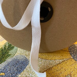 May include: A white cotton twill tape is wound around a cardboard spool. The tape is about 1 inch wide and has a slightly textured surface.