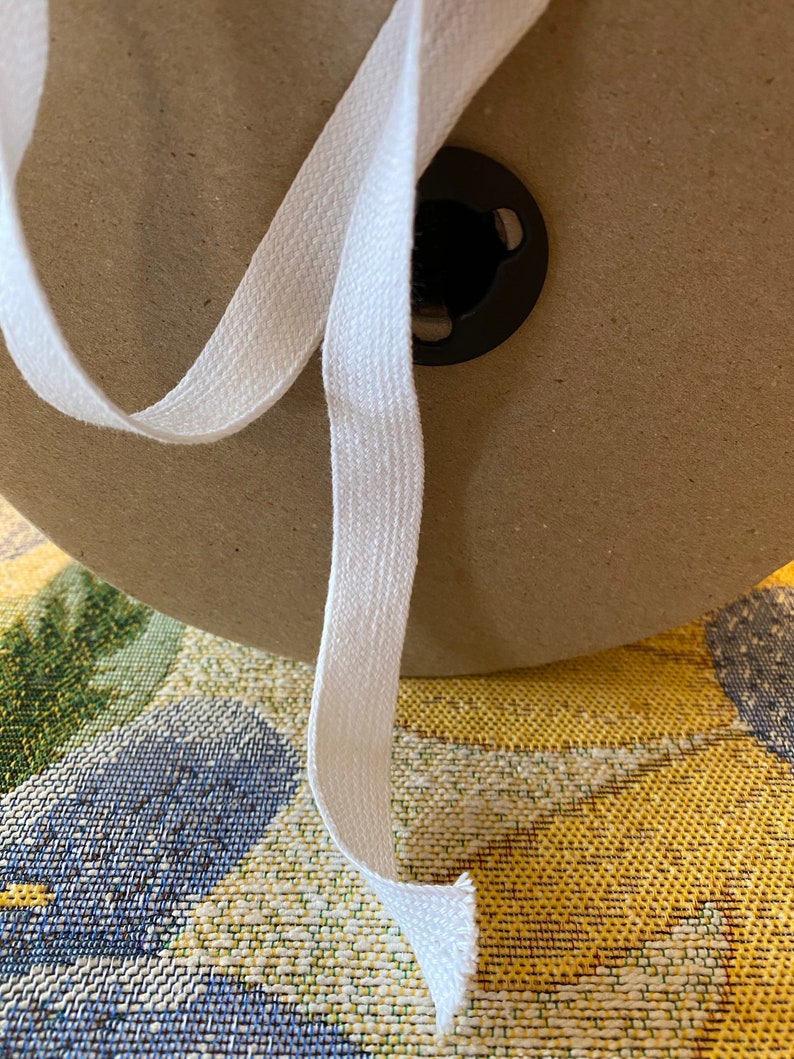 May include: A white cotton twill tape is wound around a cardboard spool. The tape is about 1 inch wide and has a slightly textured surface.