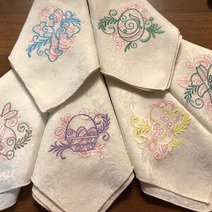 May include: Set of six white linen napkins with embroidered Easter designs. The designs include a bunny, a basket of eggs, a flower, and a swirl pattern. The napkins are perfect for adding a touch of spring to your table setting.