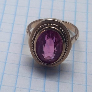 May include: A gold ring with a pink oval-shaped gemstone set in a bezel setting. The ring has a rope-like design around the bezel.