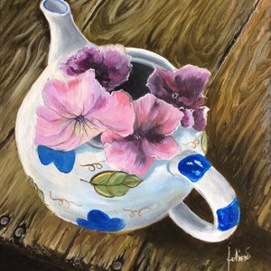 Puede incluir: Una tetera blanca con detalles azules y dorados y un diseño floral. La tetera está llena de flores rosas y moradas y está sentada sobre una superficie de madera.