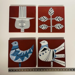 May include: Set of four square ceramic tiles with a red background. Each tile features a different design in white and blue. The designs include a stylised face with a hat, a branch with leaves, and two different birds.