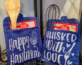 May include: Two blue oven mitts with white text and designs. One says "Happy Hanukkah" with a menorah, the other says "Whisked With Love" with a fork and hearts. Each mitt holds a Betty Crocker mix packet and a utensil.