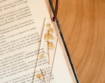 May include: A clear rectangular resin bookmark featuring a single pressed dried lily of the valley flower inside. The bookmark has a small hole at the top with a thin brown leather cord attached, resting on an open vintage book page.