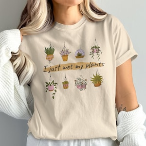 May include: Beige t-shirt featuring a botanical design with hanging plants in pots and the text "I just wet my plants" in a handwritten style. The shirt has a crew neck and short sleeves, perfect for plant lovers.