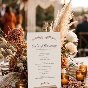 Puede incluir: Una tarjeta rectangular blanca de orden de ceremonia de boda en un soporte de madera, con el texto Order of Ceremony, Amelia & James, 14 June 2027 y una lista de eventos. Rodeada de hierba de la pampa seca, flores color óxido y velas.