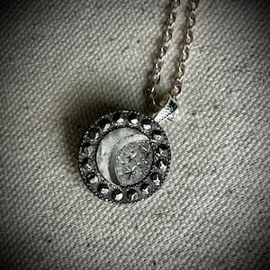 May include: Silver pendant necklace with a celestial design. The circular pendant showcases a crescent moon and stars, framed by a border of small, dark stones. The necklace has a fine chain.