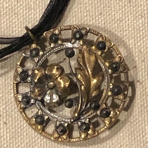 Puede incluir: Un colgante ovalado de oro y plata con un diseño floral y detalles negros. El colgante está unido a un cordón negro.