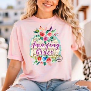 May include: Pink t-shirt with a floral design and the words "Amazing Grace" printed on it. The design includes a vintage sheet music background with lyrics from the hymn "Amazing Grace".
