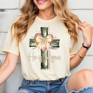 May include: A light yellow t-shirt with a green and black cross with a pink heart and gold bow. The text "He is Risen" is printed below the cross.