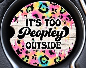 May include: A circular car cup holder coaster featuring a vibrant, multi-colored tie-dye and leopard print background over a white wood grain texture. The text IT'S TOO PEOPLEY OUTSIDE is printed in bold, black, retro-style lettering across the center.