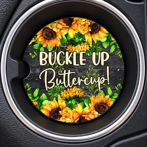 May include: A circular car cup holder coaster featuring a dark wood grain background, vibrant yellow sunflowers, and green leaves. The text BUCKLE UP Buttercup! is printed in a bold, light-colored font across the center of the coaster.