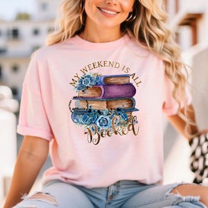 May include: A pink t-shirt with the text "My Weekend Is All Booked" in gold lettering. The text is surrounded by a stack of books with blue flowers.
