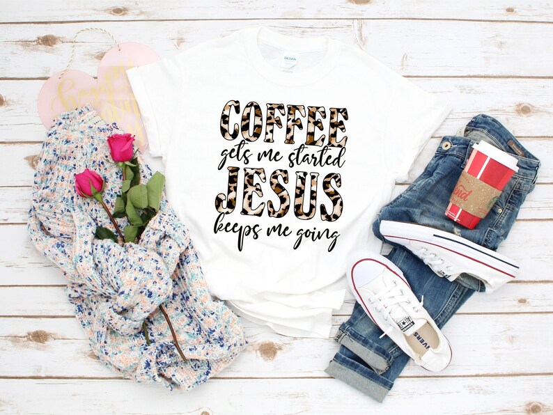 Pode incluir: Camiseta branca com um design de estampa de leopardo que diz "Coffee gets me started Jesus keeps me going". A camiseta est&aacute; colocada sobre uma superf&iacute;cie de madeira com um par de cal&ccedil;as jeans, um su&eacute;ter e uma x&iacute;cara de caf&eacute;.