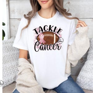 Pu&ograve; includere: Maglietta bianca con un nastro rosa e una grafica di un pallone da calcio. Il testo "Tackle Cancer" &egrave; stampato sulla maglietta.
