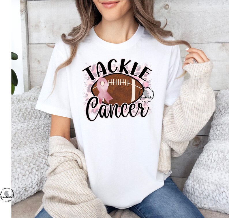 Pu&ograve; includere: Maglietta bianca con un nastro rosa e una grafica di un pallone da calcio. Il testo "Tackle Cancer" &egrave; stampato sulla maglietta.