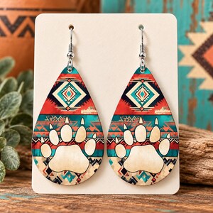 May include: A pair of teardrop-shaped earrings featuring a cream-colored bear paw print graphic centered on a background of red, teal, and black geometric patterns. The earrings hang from silver-toned metal hooks attached to a white display card.
