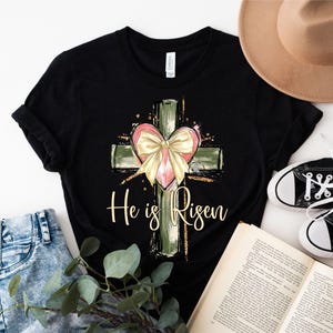 May include: Black t-shirt with a green and gold cross and a pink heart with a yellow bow. The text "He is Risen" is printed below the cross.