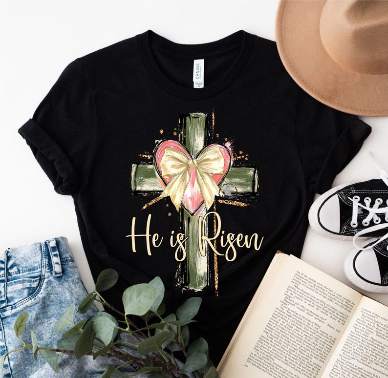 May include: Black t-shirt with a green and gold cross and a pink heart with a yellow bow. The text "He is Risen" is printed below the cross.