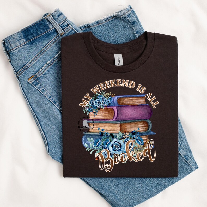 May include: A brown t-shirt with the text "My weekend is all booked" in gold lettering. The text is surrounded by a stack of books with blue flowers.