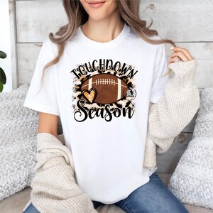 Puede incluir: Camiseta blanca con un bal&oacute;n de f&uacute;tbol marr&oacute;n y un dise&ntilde;o de estampado de leopardo. El texto "Touchdown Season" est&aacute; impreso en la camiseta.